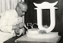 502241 Afbeelding van een banketbakker tijdens het prepareren van een taart met het logo van de Koninklijke Nederlandse ...